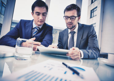 Image of two young businessmen discussing document in touchpad at meeting